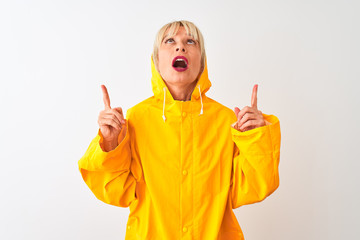 Middle age woman wearing rain coat with hood standing over isolated white background amazed and surprised looking up and pointing with fingers and raised arms.