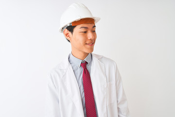 Wall Mural - Chinese architect man wearing coat and helmet standing over isolated white background looking away to side with smile on face, natural expression. Laughing confident.