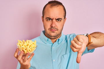 Sticker - Young man holding bowl with extruded corn standing over isolated pink background with angry face, negative sign showing dislike with thumbs down, rejection concept