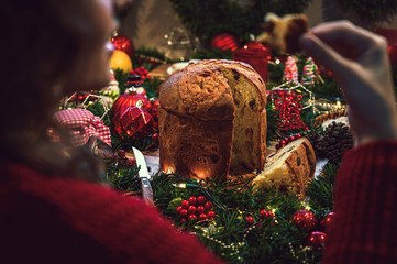 Young woman eating a piece of panettone, in the background a table of Christmas ingredients accompanied by hot chocolate, orange, cookies, cinnamon, pine.