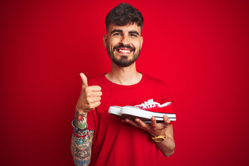 Wall Mural - Young man with tattoo holding sneakers standing over isolated red background happy with big smile doing ok sign, thumb up with fingers, excellent sign