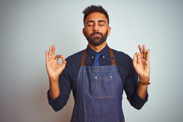 Wall Mural - Young indian employee man wearing apron uniform standing over isolated white background relax and smiling with eyes closed doing meditation gesture with fingers. Yoga concept.