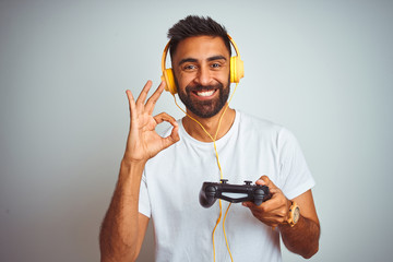 Arab indian gamer man playing video game using headphones over isolated white background doing ok sign with fingers, excellent symbol