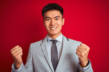 Asian chinese businessman wearing grey jacket and tie standing over isolated red background celebrating surprised and amazed for success with arms raised and open eyes. Winner concept.