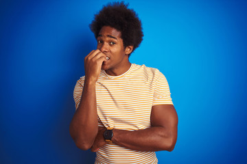 Poster - American man with afro hair wearing striped yellow t-shirt over isolated blue background looking stressed and nervous with hands on mouth biting nails. Anxiety problem.