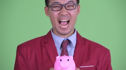 Wall Mural - Face of happy Asian businessman with eyeglasses holding piggy bank and looking surprised