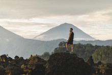 Young Man Standing On Top Of Stone And Enjoying Amazing Volcanic Mountain Agung View In Bali. Travel And Active Lifestyle Concept.
