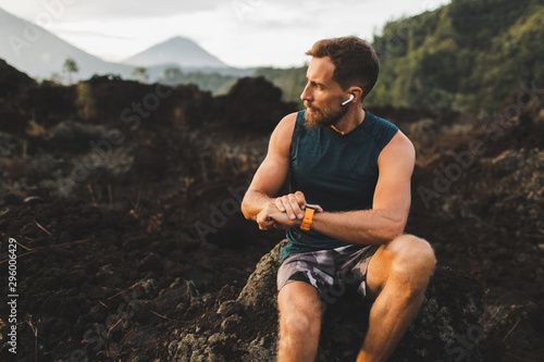 Fotografía Young hipster runner with beard using smart watch and listening music in wireless headphones before running outdoors