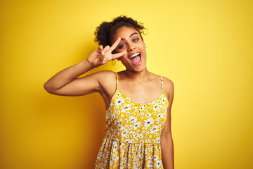 Sticker - African american woman wearing casual floral dress standing over isolated yellow background Doing peace symbol with fingers over face, smiling cheerful showing victory