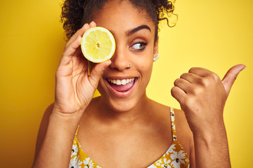 Young african american woman holding slice of lemon over isolated yellow background pointing and showing with thumb up to the side with happy face smiling