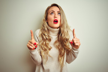 Beautiful woman wearing winter turtleneck sweater over isolated white background amazed and surprised looking up and pointing with fingers and raised arms.