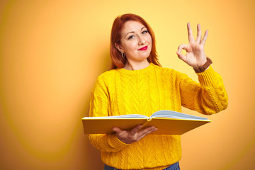 Poster - Young redhead student woman reading book standing over yellow isolated background doing ok sign with fingers, excellent symbol