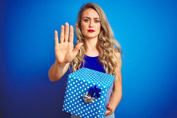 Canvas Print - Young beautiful woman holding birthday present over blue isolated background with open hand doing stop sign with serious and confident expression, defense gesture