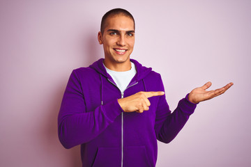 Poster - Young handsome man wearing purple sweatshirt over pink isolated background amazed and smiling to the camera while presenting with hand and pointing with finger.