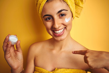 Sticker - Young woman wearing a shower towel using face cream over yellow isolated background with surprise face pointing finger to himself