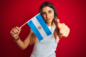 Poster - Young beautiful woman holding argentine flag over red isolated background pointing with finger to the camera and to you, hand sign, positive and confident gesture from the front