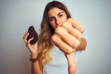 Sticker - Young beautiful woman eating avocado over grey isolated background with angry face, negative sign showing dislike with thumbs down, rejection concept