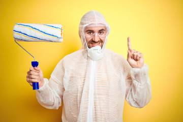 Young man wearing painter equipment and holding painting roller over isolated yellow background surprised with an idea or question pointing finger with happy face, number one