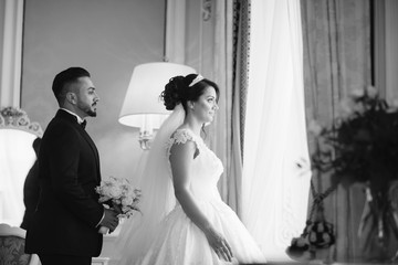 A beautiful young Turkish man wearing a black suit and a beard standing near windows along with his beautiful bride in a white dress and a bouquet of flowers