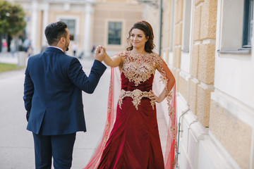 Beautiful woman in a long red dress walks around the city with her husband in a blue suit and with a beard