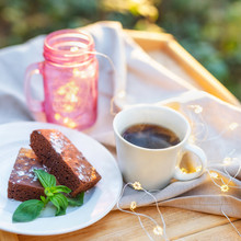 Close-up Of Cup Of Tea With A Brownie Cake Decorated With Some Mint On A Wooden Table Outdoors. Mason Jar Decorated With Lights On The Background