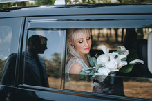 Beautiful Bride With A Bouquet In Her Hands Sitting In A Car Near The Window, Reflection Of The Groom's Silhouette. Wedding Portrait Of A Cute Blonde Girl With Flowers. Photography And Concept.