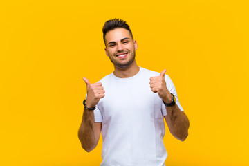 Wall Mural - young hispanic man smiling joyfully and looking happy, feeling carefree and positive with both thumbs up against orange wall