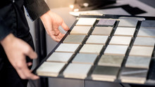 Male Architect Or Interior Designer Hand Choosing Ceramic Texture Sample From Swatch Board In Design Studio. Floor And Wall Finishing Material For Architecture And Construction Industry.