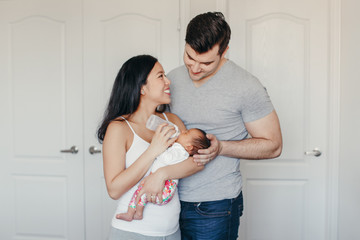 Wall Mural - Beautiful Chinese Asian mother and Caucasian father feeding newborn infant baby child from bottle with milk. Happy family in bedroom. Home lifestyle authentic moment. Positive real emotions.