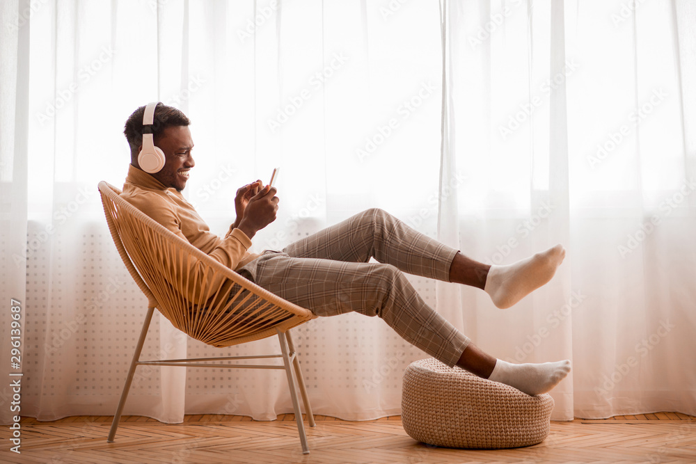Afro Guy In Headphones Using Smartphone Sitting On Chair Indoor - obrazy, fototapety, plakaty 