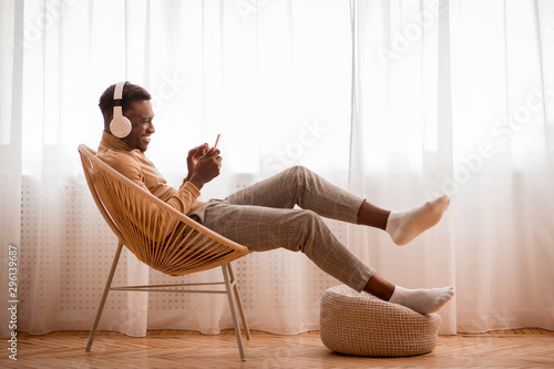 Cuadros en Lienzo Afro Guy In Headphones Using Smartphone Sitting On Chair Indoor