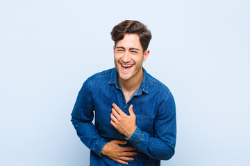 Wall Mural - young handsome man laughing out loud at some hilarious joke, feeling happy and cheerful, having fun against blue background