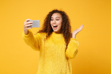 Sticker - Excited young african american girl in fur sweater posing isolated on yellow orange wall background. People lifestyle concept. Mock up copy space. Doing selfie shot on mobile phone, spreading hands.