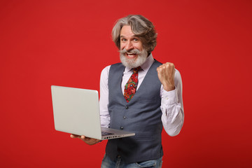 Joyful elderly gray-haired mustache bearded man in classic shirt vest tie posing isolated on red background. People lifestyle concept. Mock up copy space. Hold laptop pc computer doing winner gesture.