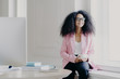 © VK Studio - Happy female entrepreneur has bushy Afro hair, wears rosy jacket and black trousers, holds cup of drink, poses at windowsill in spacious cabinet with table, modern computer, enjoys aromatic coffee