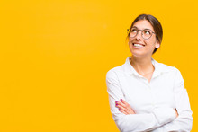 Young Businesswoman Feeling Happy, Proud And Hopeful, Wondering Or Thinking, Looking Up To Copy Space With Crossed Arms Against Orange Wall
