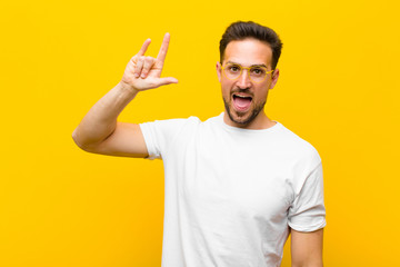 young handsome man feeling happy, fun, confident, positive and rebellious, making rock or heavy metal sign with hand against orange wall