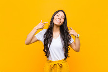 Young Pretty Latin Woman Smiling Confidently Pointing To Own Broad Smile, Positive, Relaxed, Satisfied Attitude Against Orange Wall