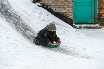 Wall Mural - Funny child dressed in adult oversized jacket riding sled down icey trek from cellar in winter day. Little girl with emotional face riding slide in the village yard.