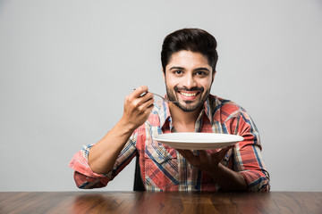 empty plate and Indian man with beard holding spoon and fork, wearing checkered shirt and sitting at table 