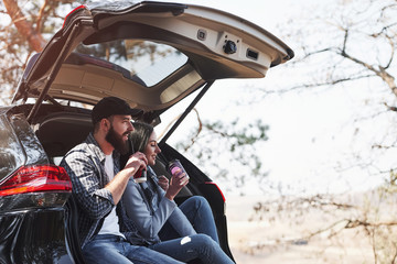 Drinking delicious coffee. Sitting on rear part of automobile. Enjoying the nature. Couple have arrived to the forest on their brand new black car