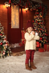 Wall Mural - cute beautiful girl in a white sweater and red hat having fun and playing snowballs in winter on the street against the background of a wooden house. nearby are Christmas trees with lights