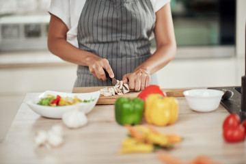 Wall Mural - Close up of mixed race woman in apron standing in kitchen and chopping mushrooms.