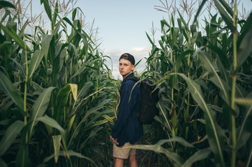 Fashionable photo of attractive young man with backpack and casual clothes standing in corn field and posing at camera. Portrait of stylish tourist guy in corn field.