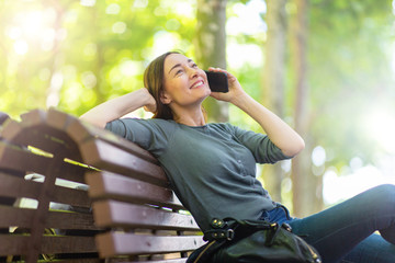 Wall Mural - happy middle aged woman sitting on park bench talking with mobile phone