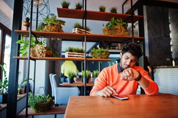 Wall Mural - Confident young indian man in smart casual wear like orange sweater, sitting at cafe and using his mobile phone.