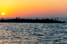 Sunset On The Mediterranean Sea. Silhouettes Of Fishermen On The Breakwaters.