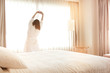 © anon - Woman stretching in bed after waking up, back view. Woman sitting near the big white window while stretching on bed after waking up with sunrise at morning, back view.