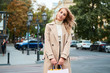 © Anton - Beautiful stylish blond girl in trench coat with shopping bags happily looking in camera on street