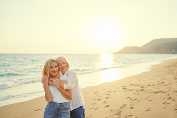 Outdoor portrait of smiling happy caucasian senior mother with her adult daughter hugging and looking at the camera on sea beach.